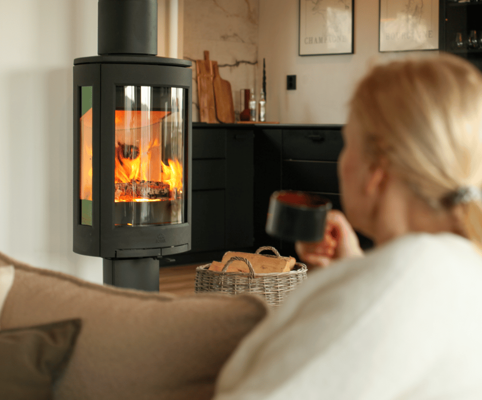 kvinne sitter i en sofa foran en vedovn Jøtul F 373 med en kopp og nyter varmen fra vedovnen